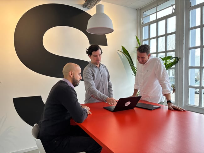 Drei QA Experten an einem roten Tisch vor einem Laptop: QA als Investionen verstehen lohnt sich