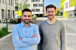 Two men stand in the courtyard between houses and smile at the camera.